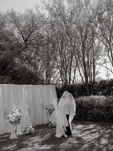Wedding kiss portrait of newlyweds sharing an under veil kiss beneath a lace veil, with garden trees and hedge softly framing them.