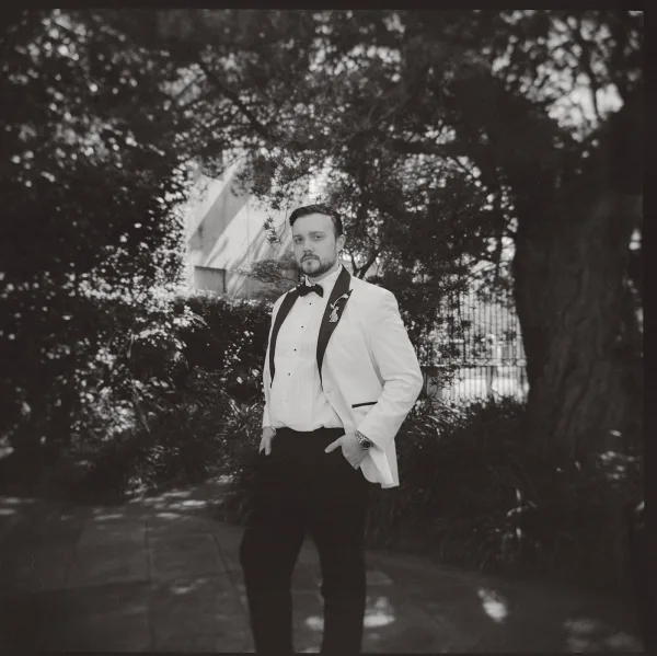 Groom portrait of a groom in a white dinner jacket standing along a garden path by an iron fence and garden trees.