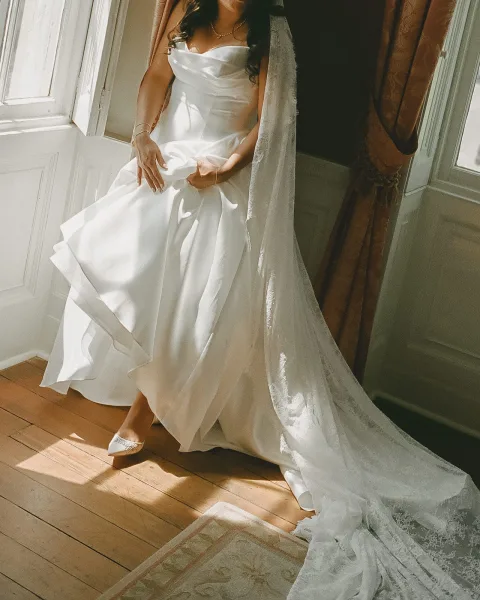 Wedding dress close-up of a strapless bridal gown, satin bodice and clean lines, paired with a long lace veil against a sunlit window.