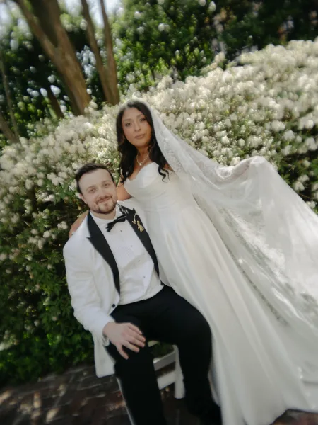 Couple portrait with bride holding veil, her lace veil trailing beside the groom amid flowering shrubs and a brick garden pathway.