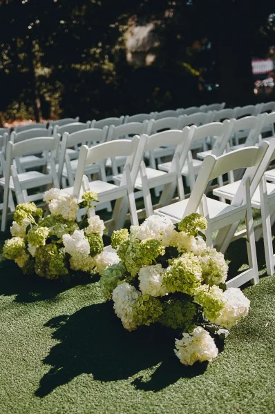 Ceremony aisle decor featuring hydrangeas, with outdoor ceremony seating along a grass lawn edged by garden trees.