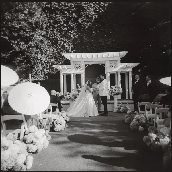Ceremony moment during the wedding vows exchange beneath a pergola altar on an outdoor aisle in a garden setting.