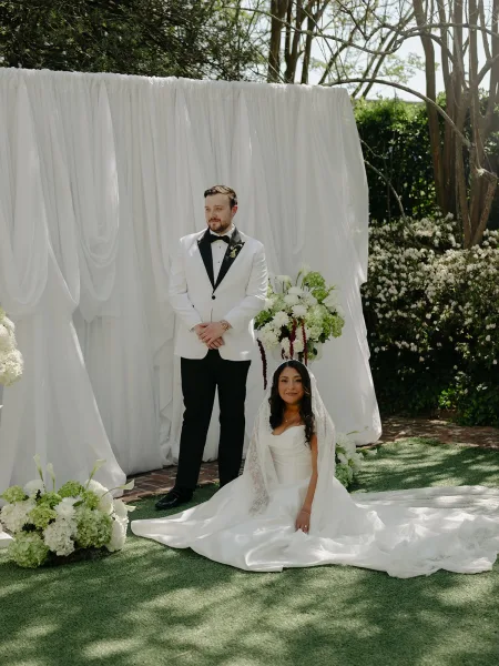 Couple portrait with a bride seated, wearing a lace veil, beside a white dinner jacket groom, against garden hedges and brick path.