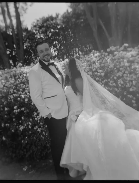 Couple portrait in an outdoor wedding portrait setting, the bride with a long veil and the groom beside her amid flowering shrubs and garden trees.
