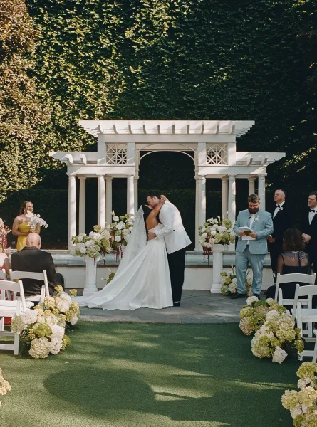Ceremony kiss, their first kiss at the altar, as the bride’s long veil trails before an ivy-covered garden wall.