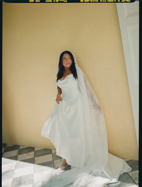 Bridal portrait of a bride in an off the shoulder wedding dress and lace veil, standing by a yellow wall and white door on a checkered tile floor.