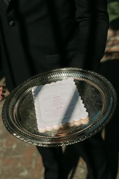 Wedding program and ceremony order card styled on a silver tray, set on a brick walkway amid garden greenery, with classic typography.