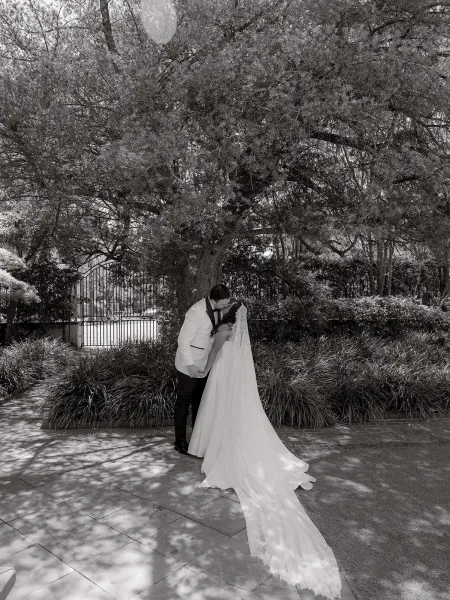 Wedding kiss portrait of newlyweds in a romantic dip kiss, her long veil flowing beside a wrought iron gate along a garden stone walkway.