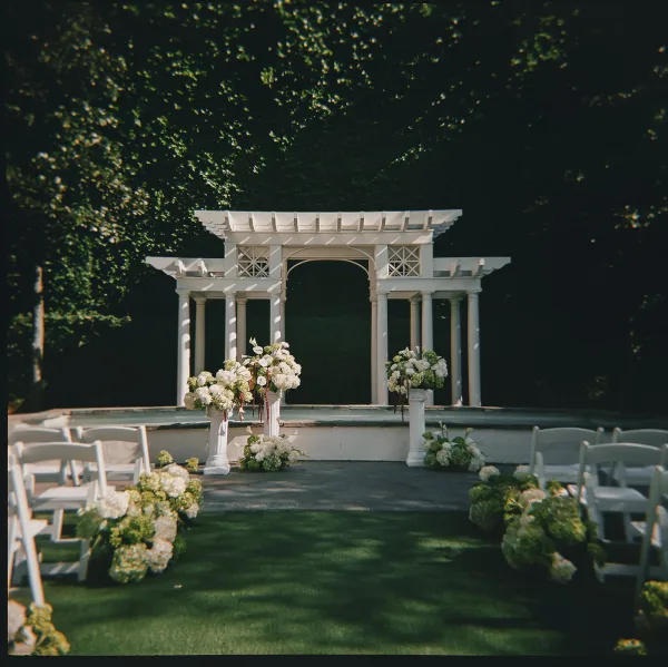 Ceremony setup with a white pergola altar, flanked by urn floral arrangements, framed by garden trees and an ivy-draped wall.