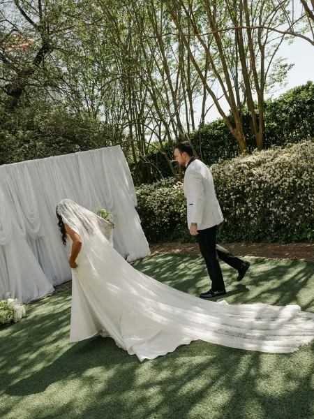 Ceremony moment with a bride with long train walking with her groom beneath draped fabric, set among garden trees and hedges along a brick path.