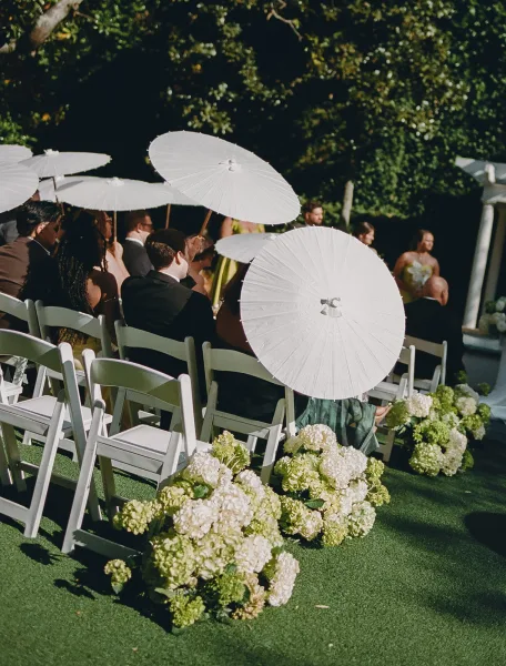 Wedding parasols arranged for guests, a thoughtful guest shade idea, on white folding chairs beneath a ceremony arbor amid garden trees and lawn.