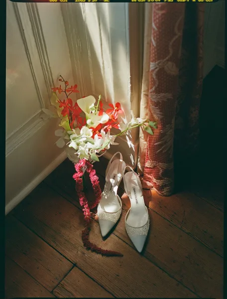 Bridal shoes—ivory lace bridal heels—rest beside an orchid bouquet on a wooden floor near a softly curtained window.