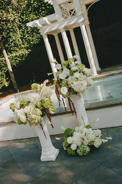 Ceremony floral arrangements as elegant pedestal flower arrangements atop white columns beneath a pergola by a reflecting pool.