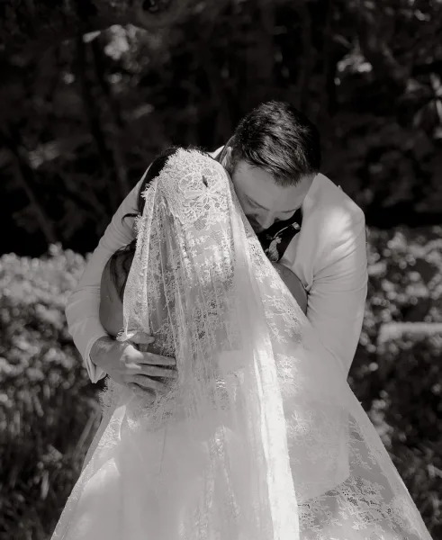 Bride and groom hug in a candid wedding moment beneath garden trees, her lace veil softly trailing as lush foliage frames their embrace.