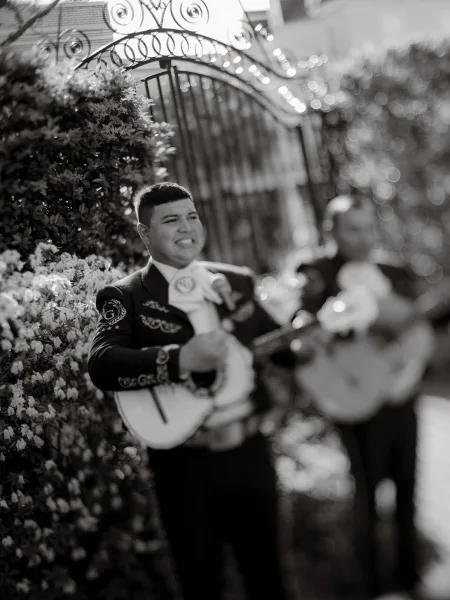 Mariachi wedding band playing wedding live music in embroidered charro suits beside a wrought iron gate and flowering shrubs in a garden.