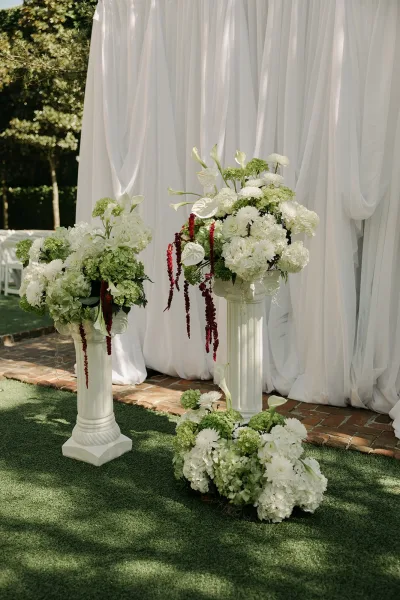 Ceremony altar decor with a draped ceremony backdrop, lush hydrangeas, and garden trees framing white chairs on a sunlit brick path.