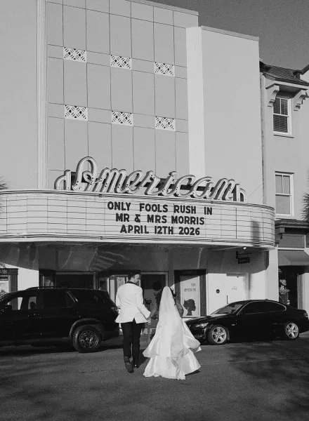 Couple portrait of a bride and groom walking hand in hand beneath a theater marquee along the street.