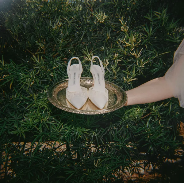 Bridal shoes—ivory wedding heels—rest on a gold tray, framed by green foliage and garden shrubs in soft light.