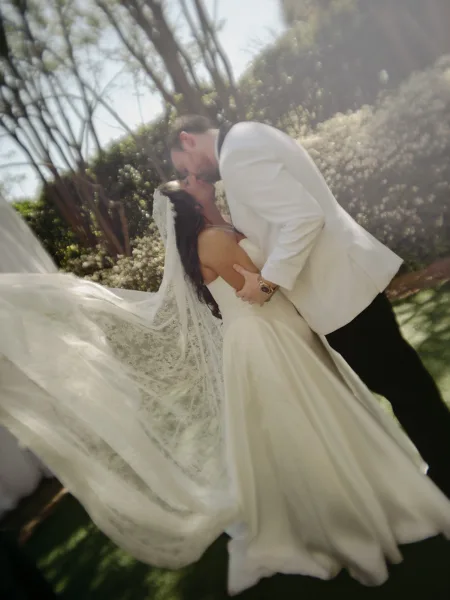 Wedding kiss portrait as a bride and groom kiss in a sunlit garden, her lace veil trailing softly while trees and hedges blur behind.