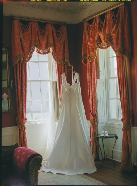 Wedding dress on hanger by tall windows with red drapery, glowing softly in a vintage room with mirror and wood floor details.