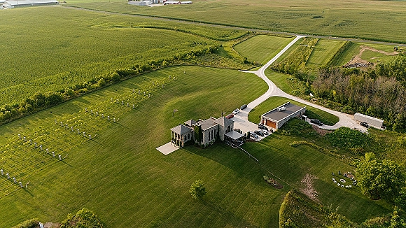 Wedding venue aerial view of a stone building with gravel driveway, parked cars, and string lights amid open farmland and trees