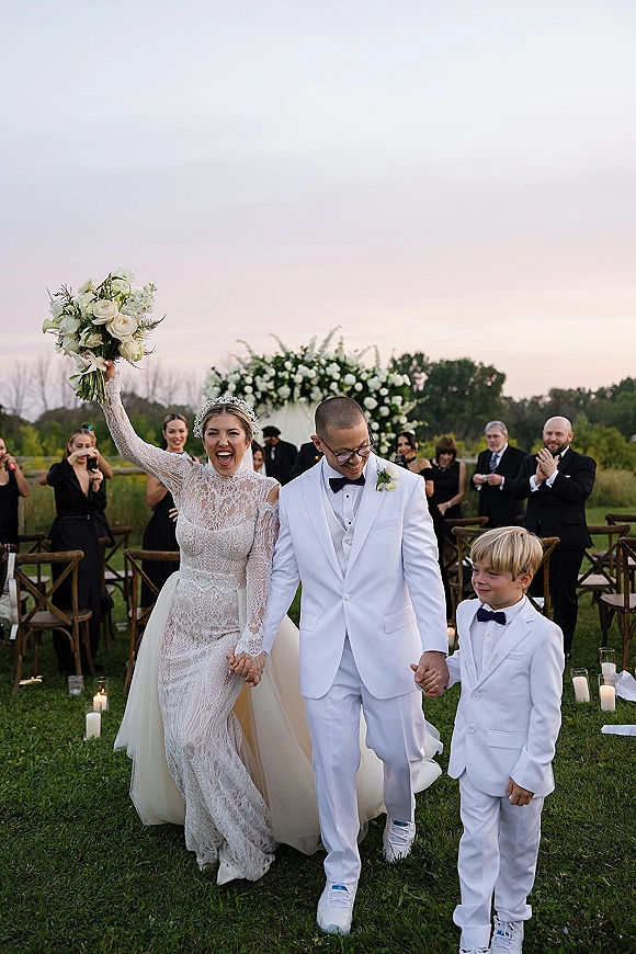 Wedding recessional with bride holding bouquet up as she and groom walk the aisle with ring bearer at a sunset lawn ceremony with guests cheering