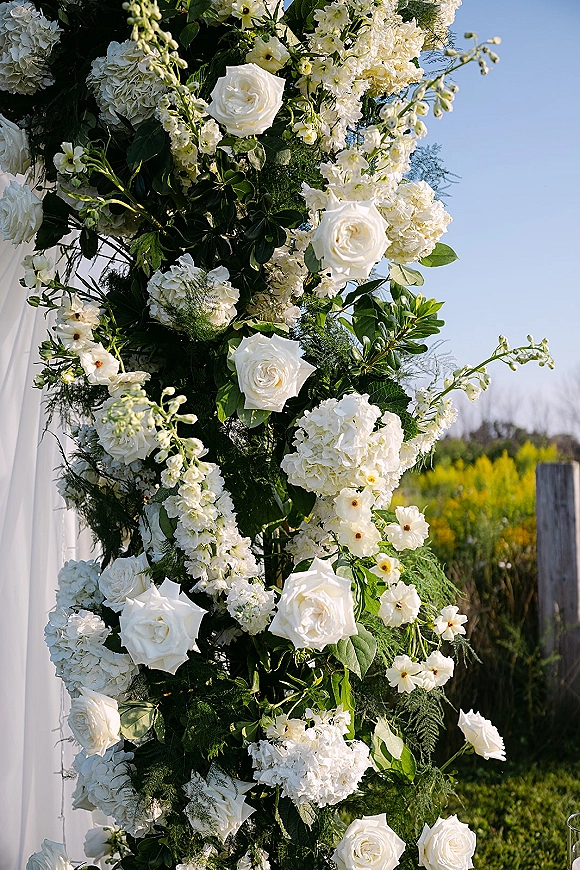 Wedding floral arch with white roses and hydrangeas, greenery and draped fabric, set against a blue sky field and wooden fence post