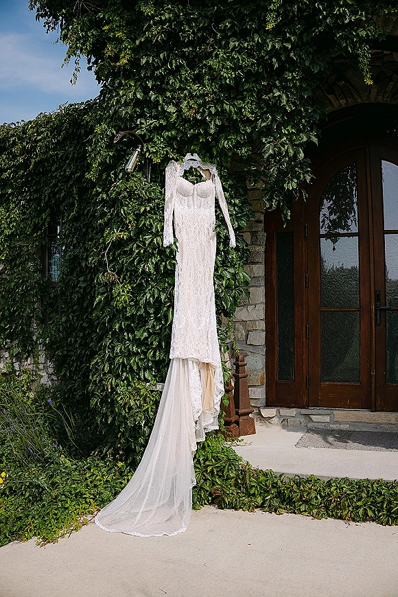 Wedding dress with lace wedding dress details hangs on a hanger, long sleeves and train against ivy-covered stone wall and arched wooden doors
