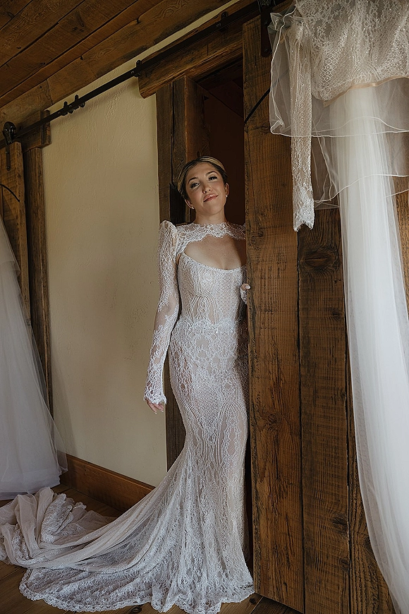 Bridal portrait in a long sleeve lace wedding dress with illusion neckline, posed by a rustic wood sliding barn door indoors