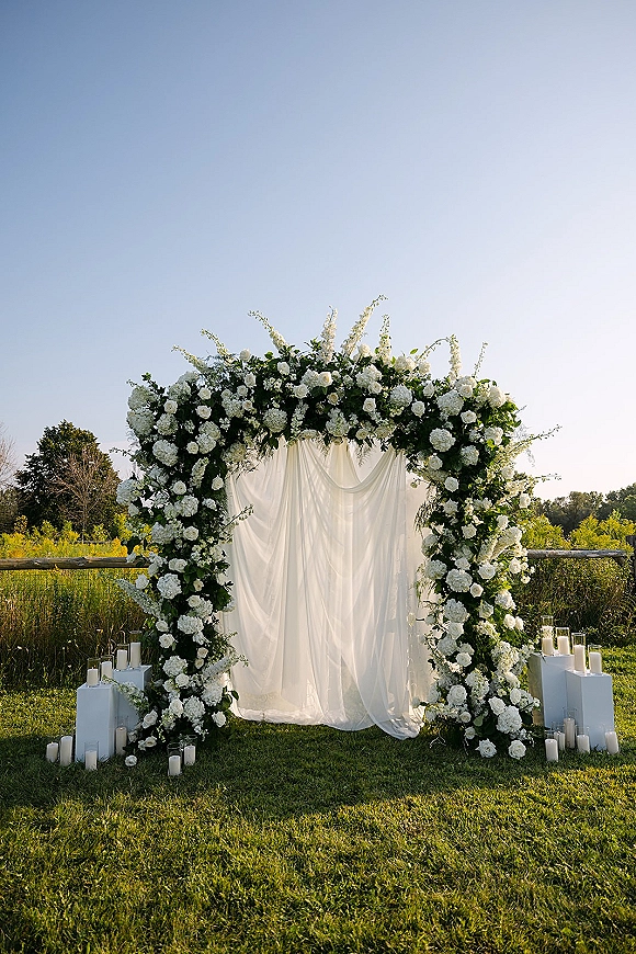 Wedding ceremony backdrop featuring a floral ceremony arch with white draping, roses, hydrangeas, greenery, and candlelit plinths on a meadow lawn