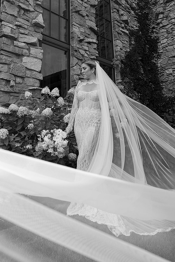 Bridal portrait in black and white of a bride in a long sleeve lace gown with cathedral veil and bouquet beside ivy stone wall and windows