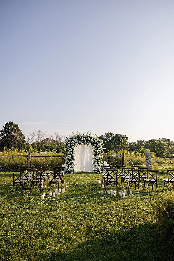 Outdoor ceremony setup with a floral arch and white drapery, candle-lined aisle, and crossback chairs on a grassy field near trees