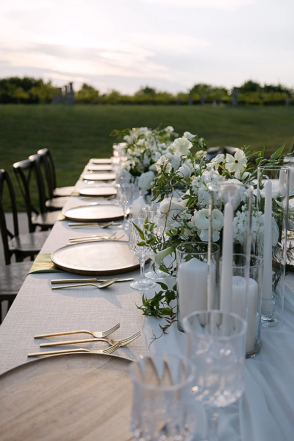 Reception tablescape with a white flower table centerpiece, greenery garland, taper candles and gold flatware on a lawn with trees beyond