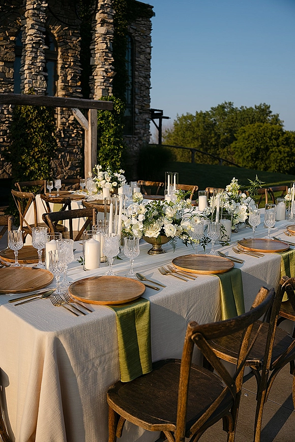 Reception tablescape with white floral centerpiece and greenery garland, gold flatware and pillar candles on a long outdoor patio table by ivy stone building