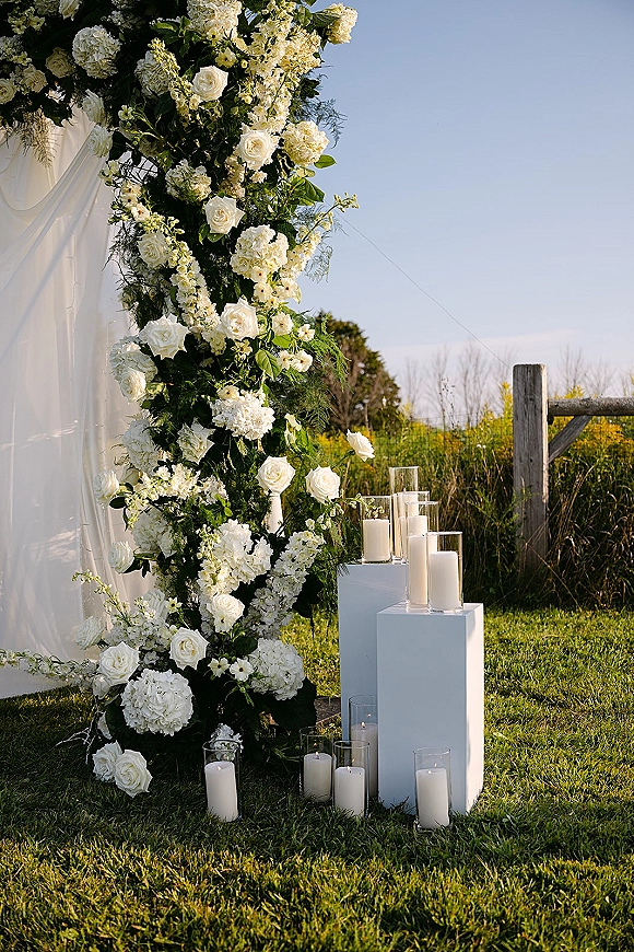 Wedding ceremony arch with a white floral ceremony arch of roses, hydrangeas, and greenery, draped fabric and candle plinths on a lawn under blue sky