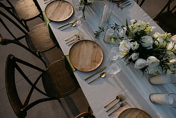Reception tablescape with wedding table runner, wood charger plates, gold flatware, clear goblets, white florals and greenery on a stone patio