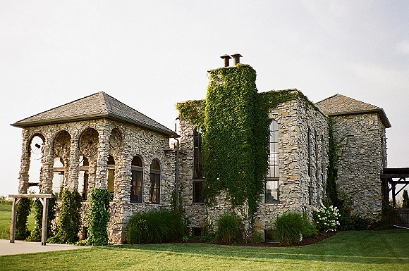 Stone wedding venue with ivy-covered walls and arched windows, framed by a pergola entrance above a manicured lawn and shrubs