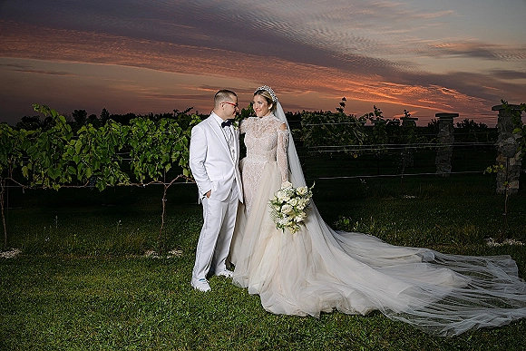 Couple portrait at sunset, bride in long sleeve lace dress with veil and white rose bouquet beside groom in white tuxedo in a vineyard