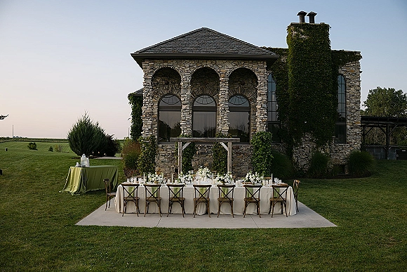 Reception tablescape with outdoor head table long banquet setup, white florals and taper candles under vine-covered pergola by ivy stone wall