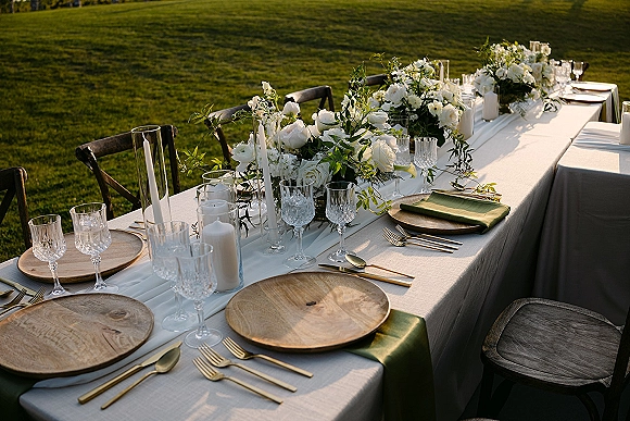 Reception tablescape with an outdoor reception table set with white floral centerpieces, greenery garland, taper candles, and crossback chairs on a grass lawn