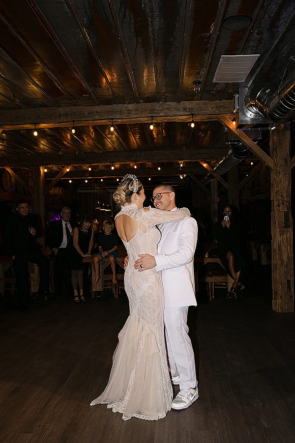 First dance of bride and groom in a lace wedding dress under string lights in a rustic barn, guests seated nearby on wood floors