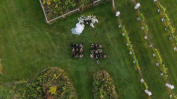 Outdoor wedding ceremony seen from above with bride and groom at a floral arch, guests seated on a white aisle fabric on a vineyard lawn