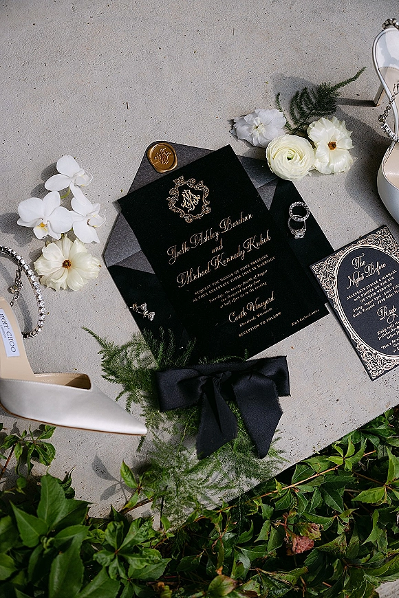 Invitation suite with black and gold wedding invitation, wax seal and envelope liner beside engagement ring, earrings, and nude heel on stone surface