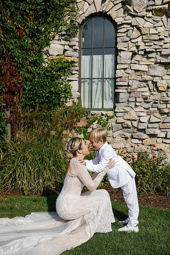 Bride and child portrait with lace wedding dress and long train, hugging a boy in a white suit and sneakers by an ivy stone wall with arched window