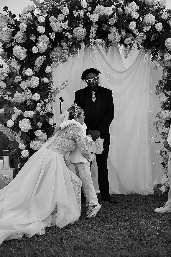 Ceremony moment as the bride hugging guest in a lace long sleeve dress and veil beneath a rose and hydrangea floral arch on a garden lawn