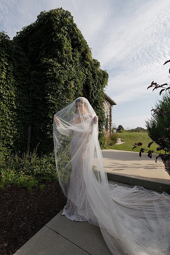 Bridal portrait of a bride holding veil in a lace wedding dress with cathedral veil and long train by an ivy-covered stone wall outdoors