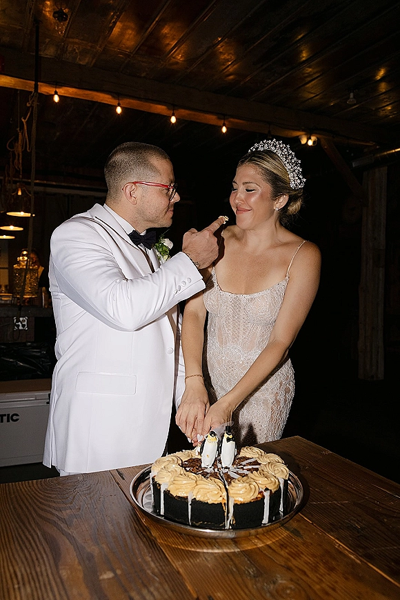 Wedding cake cutting as bride in tiara and groom in white tuxedo jacket slice a black drip cake under warm string lights in wood reception
