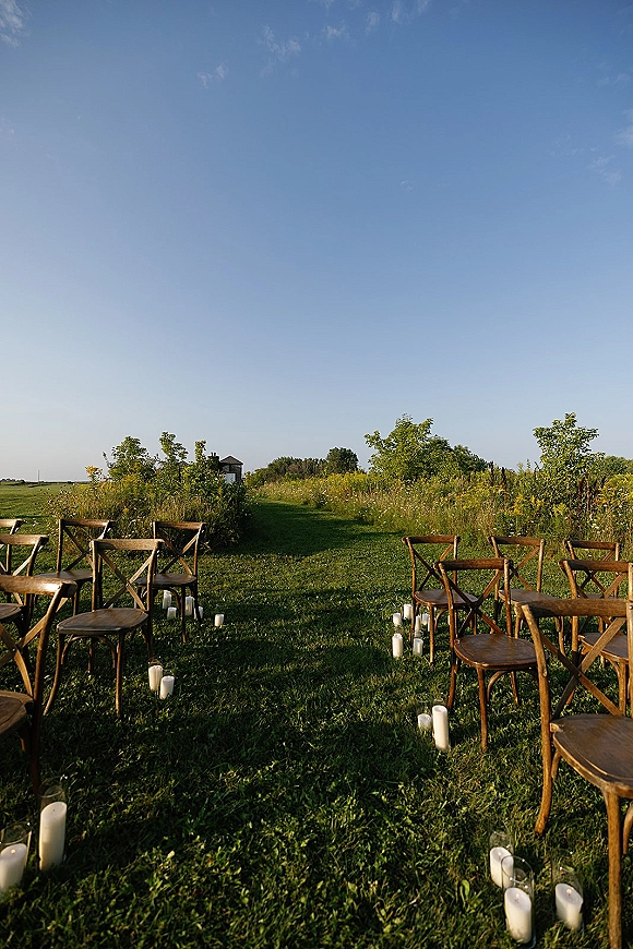 Outdoor ceremony setup with ceremony aisle chairs in two rows, candle-lined grass aisle, and wildflower meadow under blue sky