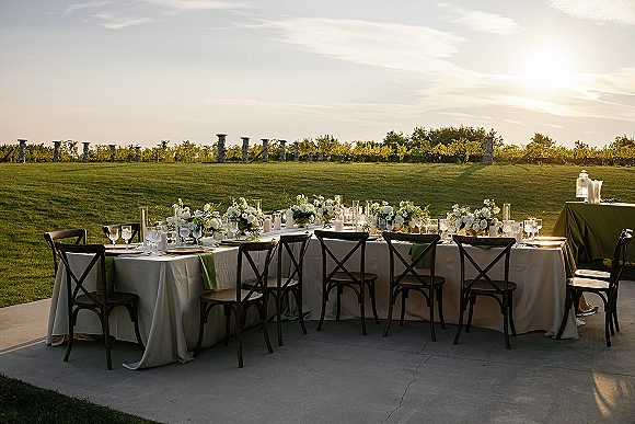 Reception tablescape with an outdoor wedding reception table set with green runner, white florals, candlelight, and vineyard rows at sunset