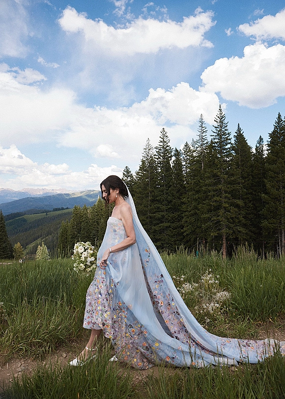 Bridal portrait of a bride in a strapless floral embroidered gown with long veil, holding a bouquet in a wildflower meadow with mountains behind